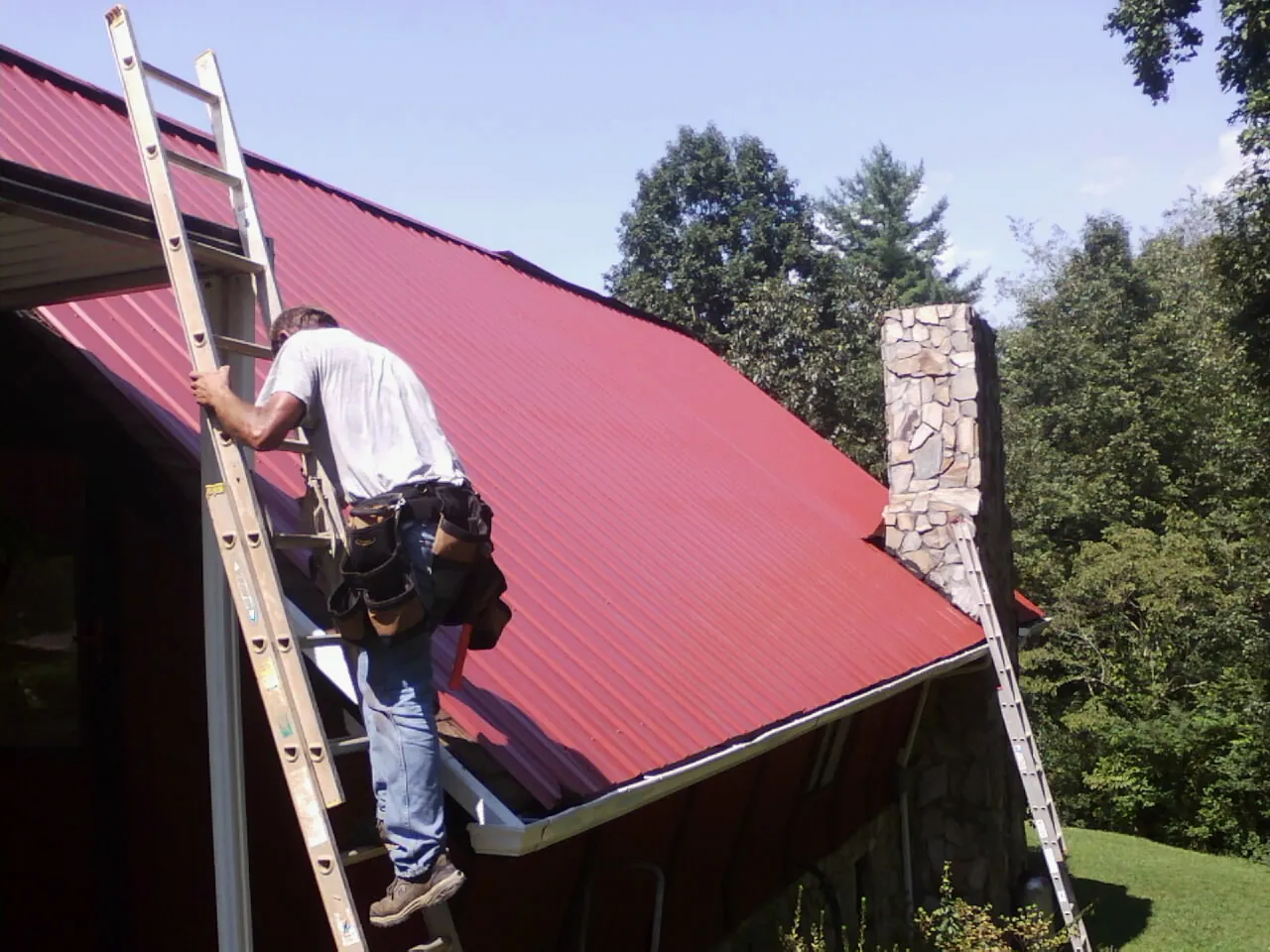 Roofer on a ladder beside a red metal roof and stone chimney — Soffit & Fascia Repair by Roofing Brothers in Phoenixville
